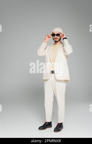 Ein trendiger schwuler Mann in beigefarbenem Anzug und Hut mit Sonnenbrille auf grauem Hintergrund, Stockbild Stockfoto
