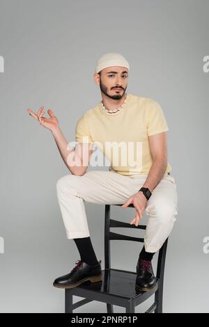 Verwirrter schwuler Mann mit Hut, der auf einem Stuhl sitzt, isoliert auf einem grauen Bild Stockfoto