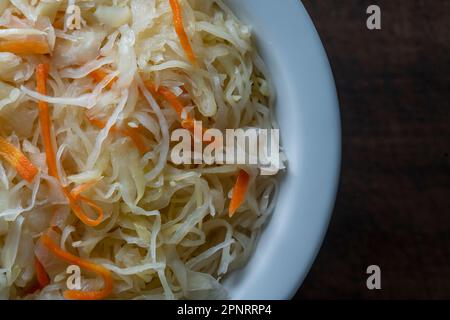 Hausgemachtes Sauerkraut in Keramikschale auf Holzhintergrund, Nahaufnahme, Draufsicht. Fein geschnittener Kohl mit Karotten, fermentiert durch Milchsäurebakterien WIT Stockfoto