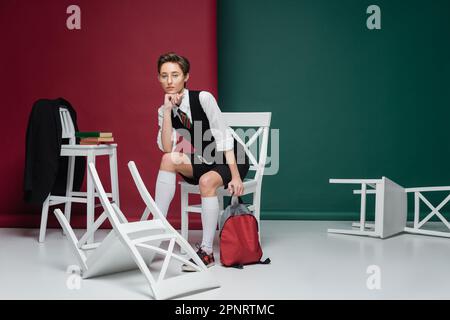 Eine lange, junge Schülerin mit Brille sitzt mit Rucksack um Stühle und Büchern auf grünem und kastanienbraunem Hintergrund, Stockbild Stockfoto