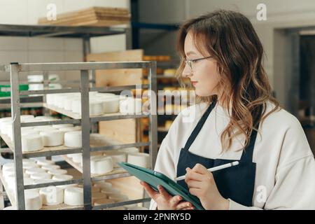 Junge Farm weibliche Besitzerin kontrolliert den Reifungsprozess von Ziegenkäseköpfen, die in den Regalen des Lagerhauses der Käserei platziert werden. Notizen mit Digital T schreiben Stockfoto