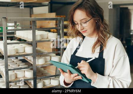 Junge Farm weibliche Besitzerin kontrolliert den Reifungsprozess von Ziegenkäseköpfen, die in den Regalen des Lagerhauses der Käserei platziert werden. Notizen mit Digital T schreiben Stockfoto