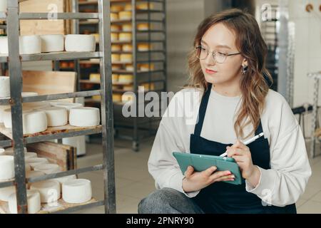 Junge Farm weibliche Besitzerin kontrolliert den Reifungsprozess von Ziegenkäseköpfen, die in den Regalen des Lagerhauses der Käserei platziert werden. Notizen mit Digital T schreiben Stockfoto