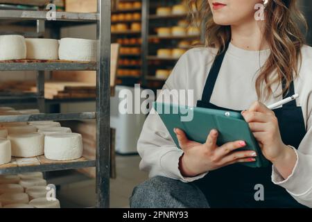 Junge Farm weibliche Besitzerin kontrolliert den Reifungsprozess von Ziegenkäseköpfen, die in den Regalen des Lagerhauses der Käserei platziert werden. Notizen mit Digital T schreiben Stockfoto