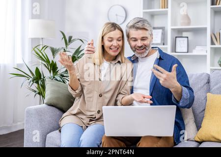 Ein reifes Ehepaar, Mann und Frau sitzen auf dem Sofa, unterhalten sich mit Freunden per Videoanruf, sitzen auf dem Sofa im Wohnzimmer, benutzen einen Laptop und lächeln fröhlich. Stockfoto