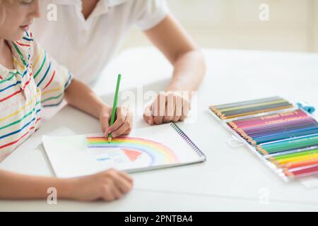 Kindzeichnung. Ein Schulkind malt ein Bild. Ein kleiner Junge mit bunten Bleistiften macht Hausaufgaben in einem weißen, sonnigen Raum. Kinder zeichnen und malen. Stockfoto