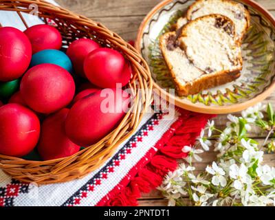 Rote Ostereier neben blühenden Zweigen und Teller mit süßem Schwammkuchen oder Kokon auf einem Holztisch Stockfoto