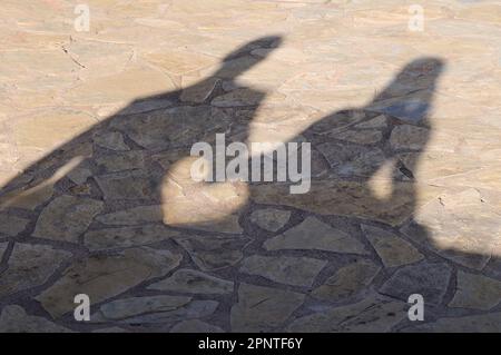 Schatten auf dem Steinboden eines Paares, das an einer Wand sitzt und Hände hält und ein Herz zwischen den Händen schafft Stockfoto