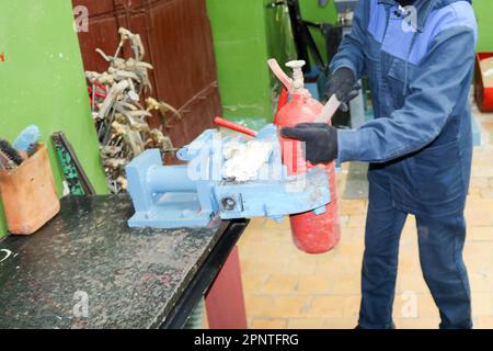 Ein männlicher Arbeiter auf einem großen industriellen Schraubstock aus Metall repariert einen roten Feuerlöscherzylinder in einer Werkstatt im Werk. Stockfoto