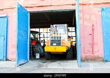 Großer, leistungsstarker gelber Traktor mit großen Rädern im Hangar, Lagerhaus, Garage mit offenen Toren. Stockfoto