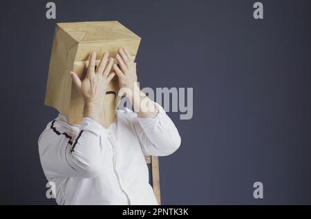 Ein trauriger Mann in einem weißen Hemd mit einer Tasche auf dem Kopf, mit einem gezeichneten traurigen Emoticon, hat Angst und schließt seine Augen mit seinen Händen. Emotionen und Gesten. Stockfoto