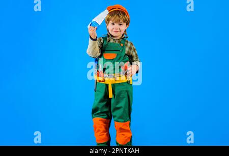 Baumeister mit Schutzhelm und Werkzeuggürtel mit Säge. Kind in Baukleidung mit Werkzeuggürtel. Bauwerkzeuge. Kleiner Reparaturtechniker mit Säge. Arbeit Stockfoto