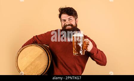 Fröhliche Brauerin mit Becher Bier und Holzfass. Ausrüstung zur Bierzubereitung. Brauereikonzept. Ein hübscher bärtiger Mann, der Bier trinkt. Ein Mann, der den Zug probiert Stockfoto