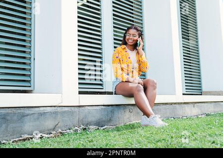 Junge Studentin, die einen Anruf auf dem Handy entgegennimmt. Eine junge afroamerikanische Teenagerin, die mit einem Mobiltelefon spricht und sich draußen auf dem Universitätscampus freundlich unterhält. Hochwertiges Foto Stockfoto