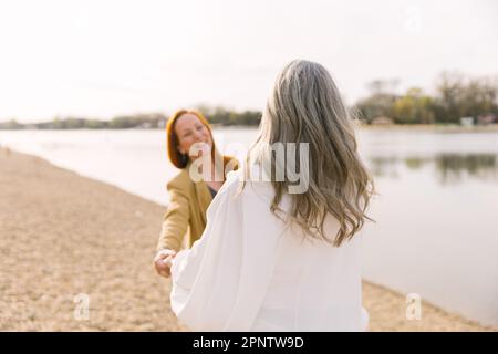 Eine Erwachsene Tochter mit einer grauhaarigen Mutter auf einem Spaziergang am Strand, lachend und lächelnd. Glückliches Alter und Altenpflege Stockfoto
