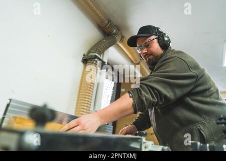 Zimmermann bei der Arbeit. Fokussierter Holzarbeiter in Schutzausrüstung mit Tischsäge. Ansicht aus niedrigem Winkel. Hochwertiges Foto Stockfoto