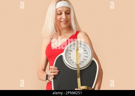 Sportliche Erwachsene Frau mit Waage und Maßband auf beigefarbenem Hintergrund Stockfoto