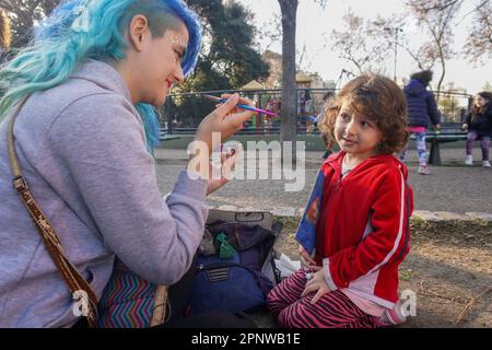 Magalí Murúa bereitet sich darauf vor, am 22. Juli 2022 in einem Park in Buenos Aires, Argentinien, Glitzer auf die Wange von Lola Ferreyra Puchatt, 4, zu kleben. Seit sie 2020 ihren Job verloren hat, hat Murúa bei Protesten, Parks und Veranstaltungen Gesichter gemalt. Die Preise für ihre Dienstleistungen sind gleitend, sodass sie für alle erschwinglich sind. (Lucila Pellettieri/Global Press Journal) Stockfoto