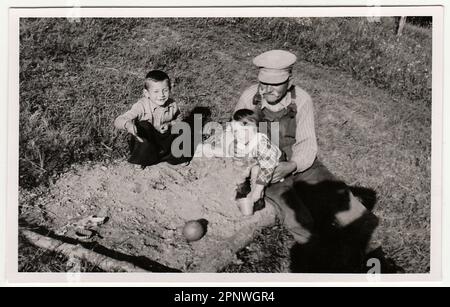 Ein klassisches Foto zeigt, wie Großvater mit Kindern im Sandkasten spielt. Antikes Schwarz-Weiß-Foto zeigt den Schatten eines Fotografen. Stockfoto