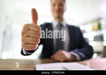Glücklicher Geschäftsmann zeigt Daumen-hoch-sitzen am Schreibtisch im Büro Stockfoto