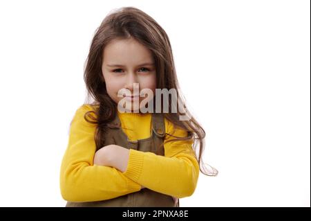 Ein bezauberndes Mädchen, das fröhlich vor der Kamera lächelt, die Arme zusammengeklappt hält, Freude und Stolz für großartige Ergebnisse in der Schule hat Stockfoto