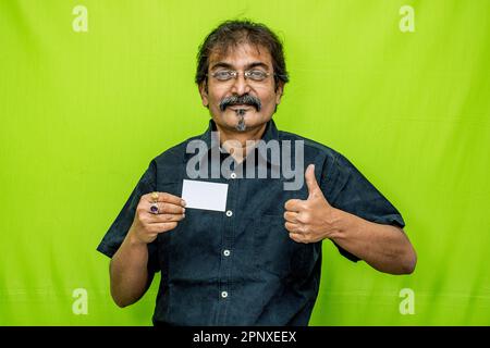 Ein Geschäftsmann in einem schwarzen Hemd und einer schwarzen Brille steht vor einem grünen Hintergrund, lächelt und hält eine Kredit-/Debitkarte in der rechten Hand Stockfoto