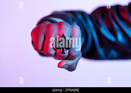 Der junge schwarze Kerl zeigt seinen großen Ring mit Pik-Symbol, das seine Faust in einer Nahaufnahme in einer roten Seitenbeleuchtung über Weiß ballt Stockfoto