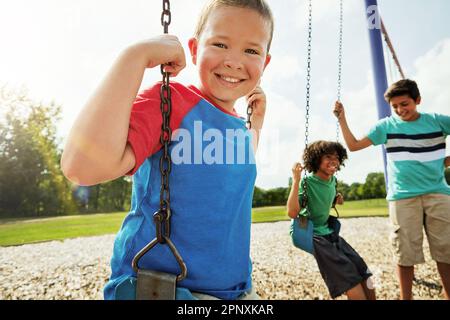 Ich will meine Schaukelkünste sehen. Porträt eines Jungen, der mit seinen Freunden auf einer Schaukel im Park spielt. Stockfoto