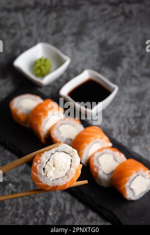 Auswahl an frischen, leckeren Sushi-Brötchen mit Lachs und Käse. Stäbchen mit Sushi auf dunklem Hintergrund. Wasabi und Soja Stockfoto