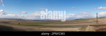 Landwirtschaftliche gepflügte Felder vor dem Hintergrund einer Bergkette und einem blauen Himmel mit weißen Wolken. Stromleitung. Friedliche Landschaft Stockfoto