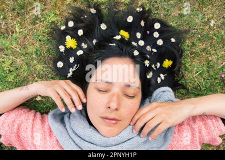 Eine Frau, die auf dem Rasen liegt, mit Blumen im Haar, Augen geschlossen und entspannt. Konzept der Frühlingsentspannung und Verbindung mit der Natur. Draufsicht. Stockfoto
