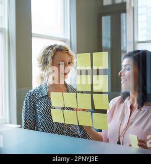 Sie sind voller großartiger Ideen. Zwei Geschäftsfrauen, die in einem Büro mit Haftnotizen an einer Glaswand Brainstorming betreiben. Stockfoto