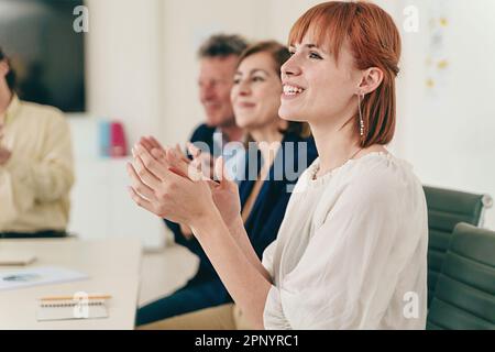 Eine Gruppe von lässig gekleideten Kollegen applaudiert am Schreibtisch, mit einer jungen rothaarigen Frau im Vordergrund und zwei leitenden Kollegen in Stockfoto