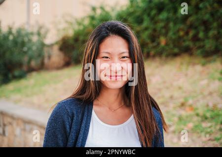 Nahaufnahme individuelles Porträt einer jungen chinesischen Frau, die lächelt und auf die Kamera schaut. Vorderansicht einer asiatischen Teenagerin mit freundlicher Einstellung. Kopfschuss einer positiven Frau, die draußen steht. Hochwertiges Foto Stockfoto