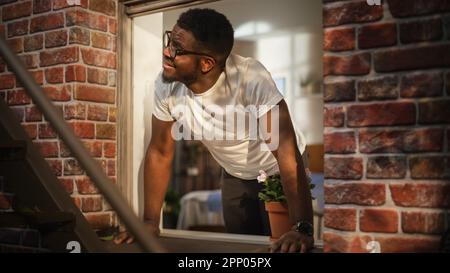 Der Schwarze öffnet morgens sein Schlafzimmerfenster und füllt seine Lungen mit frischer Luft. Happy Young Male zog in eine neue Stadt und genoss seine Wohnung. Stockfoto