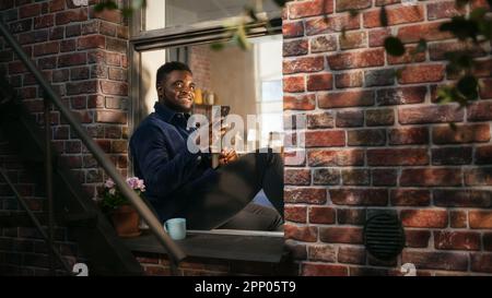 Schwarzer authentischer Mann, der sein Smartphone in seiner Wohnung benutzt, während er auf der Fensterbank des Schlafzimmers sitzt. Afroamerikanischer Mann, der Fotos in sozialen Medien postet und verwendet Stockfoto