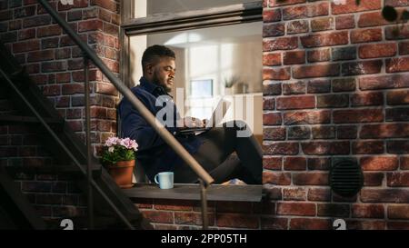 Schwarzer junger Mann lächelt und verwendet Laptop, während er auf einer Fensterbank sitzt. Afroamerikanischer Geschäftsmann genießt das Wetter, während er von zu Hause aus arbeitet. Stockfoto