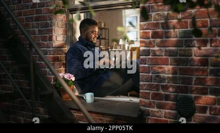 Schwarzer junger Mann lächelt und verwendet Laptop, während er auf einer Fensterbank sitzt. Afroamerikanischer Geschäftsmann genießt das Wetter, während er von zu Hause aus arbeitet. Stockfoto