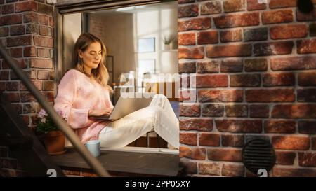 Beautiful Woman in Pink Sweater lächelt, tippt auf dem Laptop und sitzt auf ihrer Fensterbank. Weibliche Bloggerin schreibt einen Artikel über Wohlbefinden Stockfoto