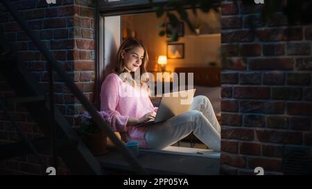 Weiße junge Frau, die nachts mit einem Notebook von Comfort of her Home auf einer Fensterbank sitzt. Weibliche Kundenberaterin, Die Dem Kundendienst Antwortet Stockfoto