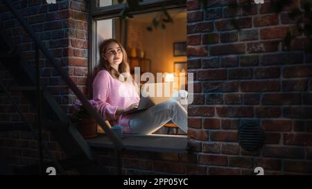 Weiße junge Frau, die nachts mit einem Notebook von Comfort of her Home auf einer Fensterbank sitzt. Weibliche Kundenberaterin, Die Dem Kundendienst Antwortet Stockfoto