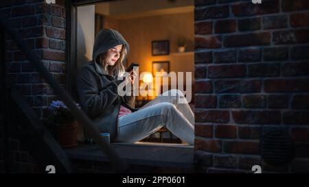 Eine weiße Frau trägt einen Hoodie mit ihrem Smartphone, während sie nachts auf einer Fensterbank sitzt. Weibliche Gamer, Die Süchtig Machende Mobiltelefonspiele Spielen Stockfoto