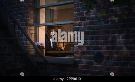 Porträt eines jungen schwarzen kreativen Mannes, der nachts von zu Hause aus auf einem Laptop in einem gemütlichen Apartment arbeitet. Afroamerikanischer Männlicher Entwickler Stockfoto