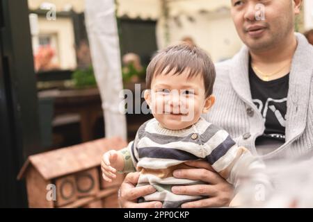 Smiley männlicher Säugling, der auf den Beinen seines Vaters sitzt und glücklich in die Kamera schaut, Dad hält das Baby mit seinen Händen. Sitzen im Freien in einem Biergarten Stockfoto