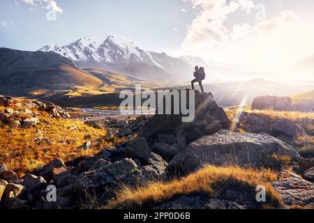 Small man Tourist mit großem Rucksack ist Wandern Klettern im Bergtal mit verschneiten Gipfeln bei Sonnenuntergang bewölkter Himmel Hintergrund in Zentralasien, Kasachst Stockfoto