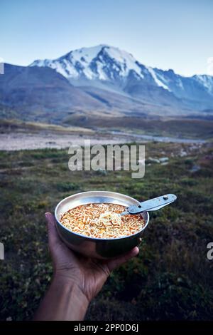 Touristen, die eine Schüssel voller Nudeln, Fast Food und einen Löffel in der Hand halten, in der Natur Bergtal Camping auf einer Wanderung durch Zentralasien, Kasachstan Stockfoto