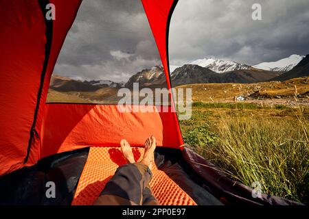 Blick aus dem Inneren eines Zelts und Wanderbeine in das Gebirgstal mit Gletscherlandschaft, pov-Blick Trekking in Zentralasien Kasachstan, Almaty Stockfoto