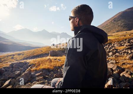 Porträt junger Mann Wandertourist in schwarzer Jacke, sitzt auf dem Felsen vor dem wunderschönen Sonnenuntergang im Bergtal. Outdoor- und Trekking-Konzept. Stockfoto