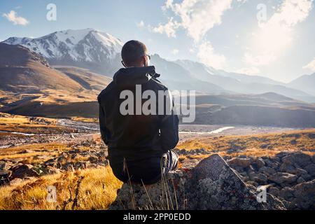 Porträt junger Mann Wandertourist in schwarzer Jacke, sitzt auf dem Felsen vor dem wunderschönen Sonnenuntergang im Bergtal. Outdoor- und Trekking-Konzept. Stockfoto