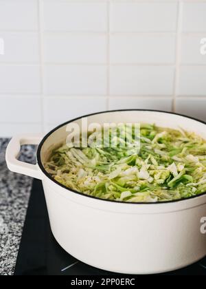 Weiße Keramik-Bratpfanne mit gehacktem Kohl in Brühe auf dem Herd. Stadium der Herstellung von Gemüsesuppen Stockfoto